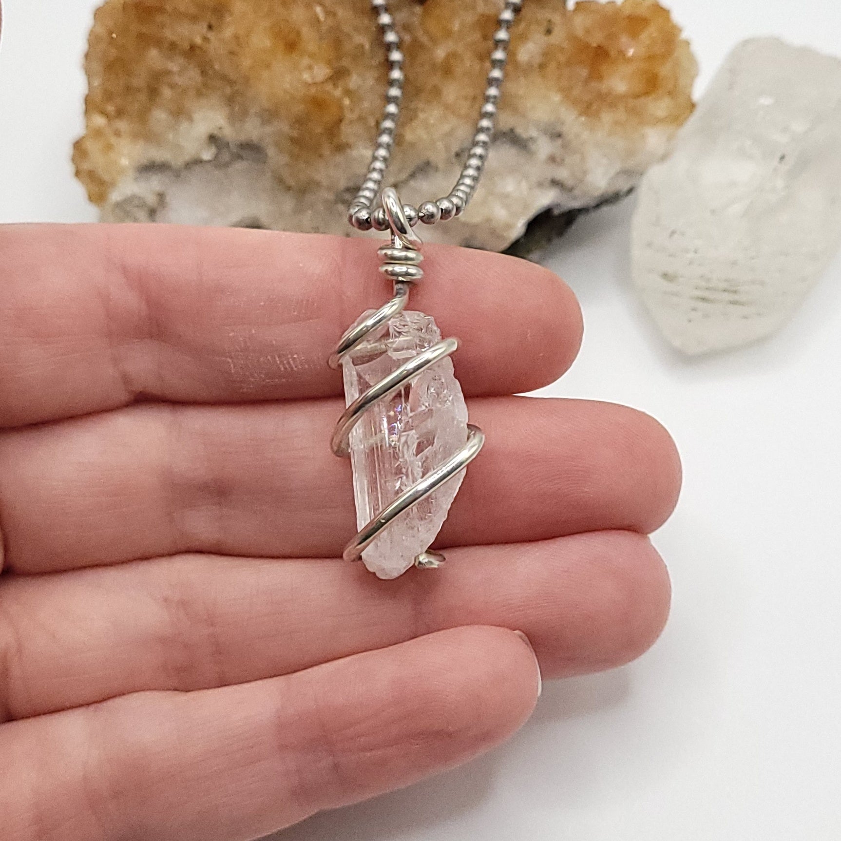 A silver-wired raw Danburite crystal necklace with a stainless-steel ball chain, held in a person's hand, with crystal stones and a resin rock on the background.