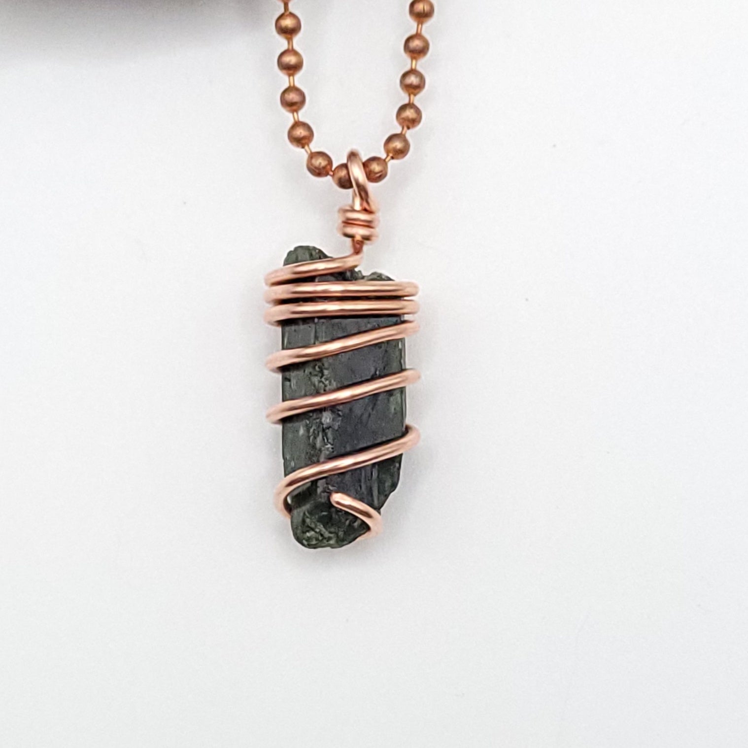 A raw Diopside crystal wrapped in copper wire with a copper ball chain close up on white background