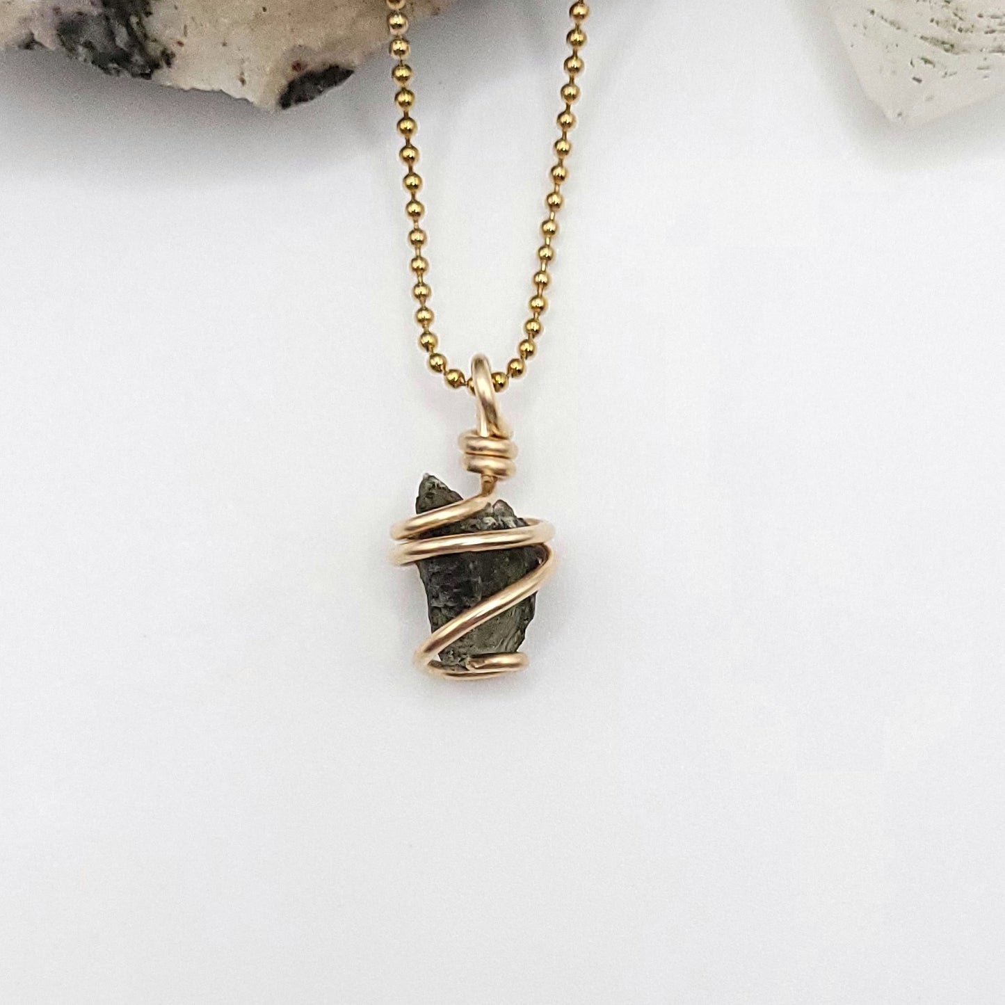 A raw Moldavite crystal pendant wrapped in gold filled wire on a gold ball chain, displayed against a white background.
