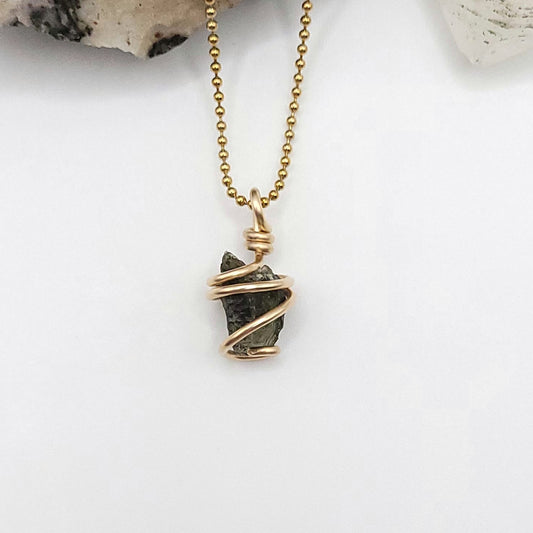 A raw Moldavite crystal pendant wrapped in gold filled wire on a gold ball chain, displayed against a white background.