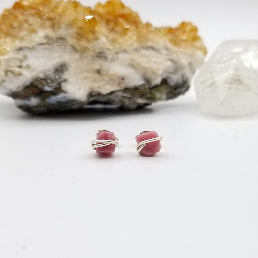 A pair of sterling silver wire wrapped rhodonite crystal stud earrings, displayed against a natural stone background.
